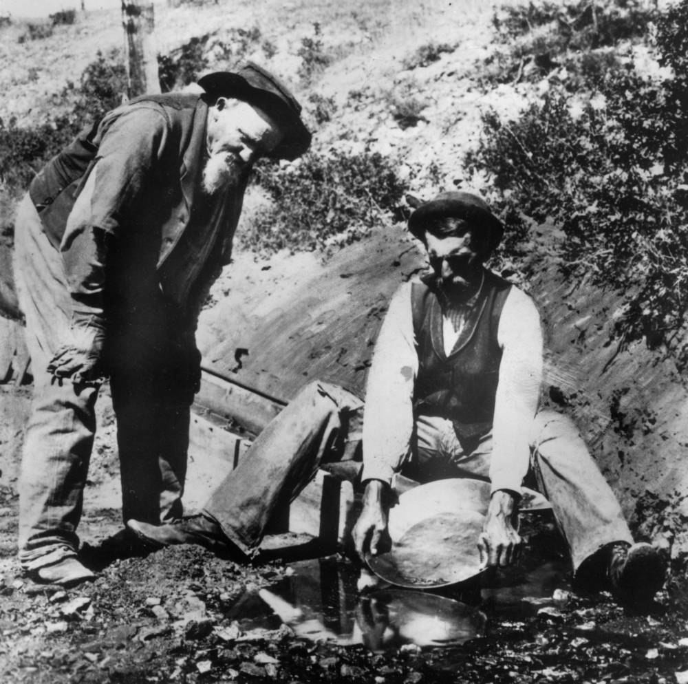 	
            Men pose near a sluice in Nelson Gulch, Lewis and Clark County, Montana. One man pans for gold, another looks on. 